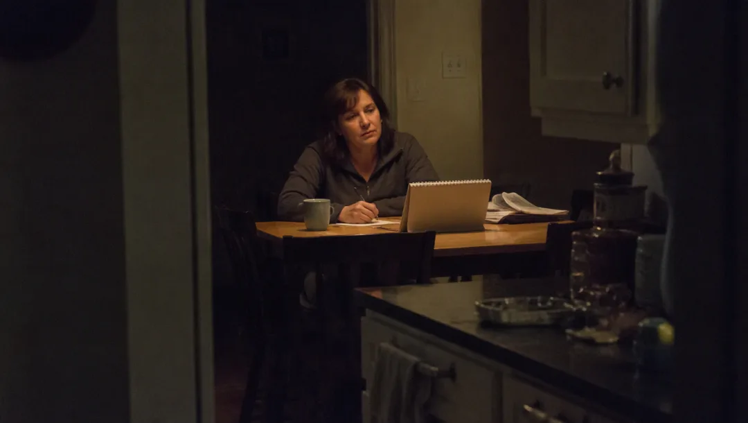 A woman alone at her kitchen table late at night, pen stopped mid-count, looking past her notebook into the middle distance