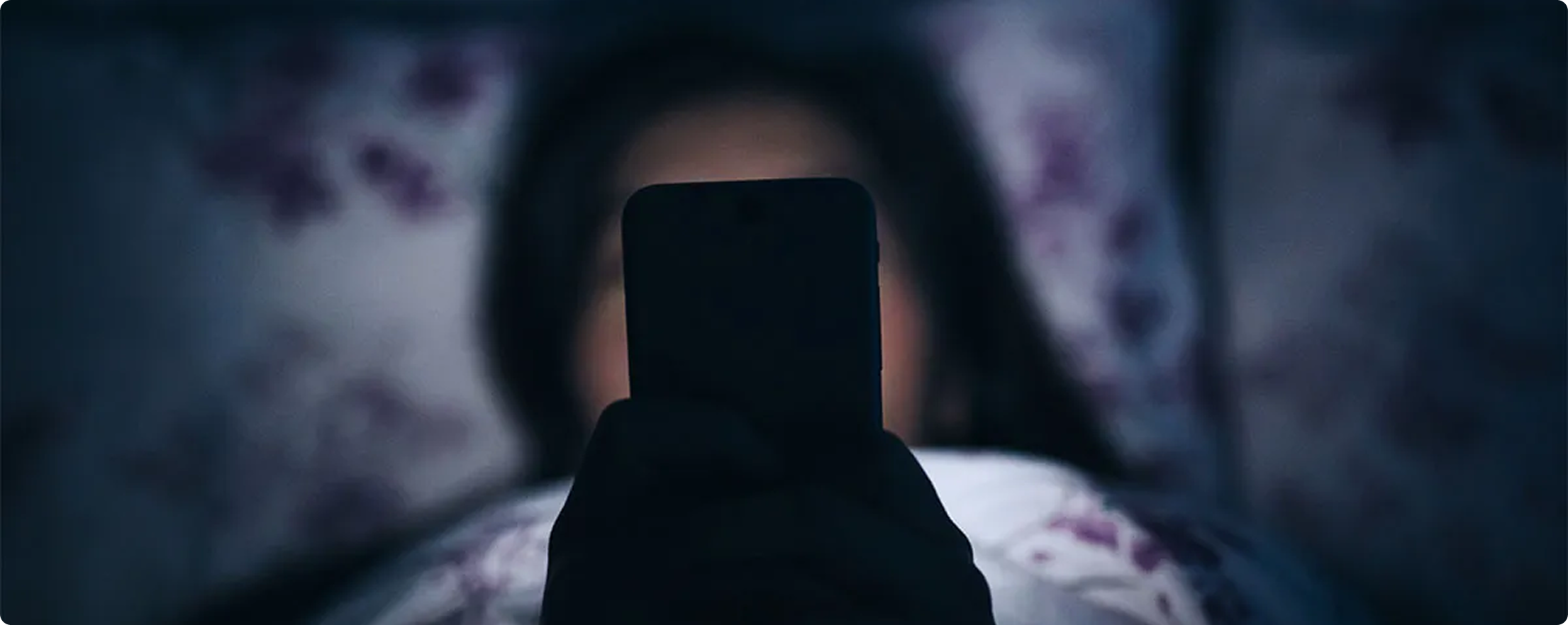 Person holding a smartphone in a dimly lit room with floral curtains.